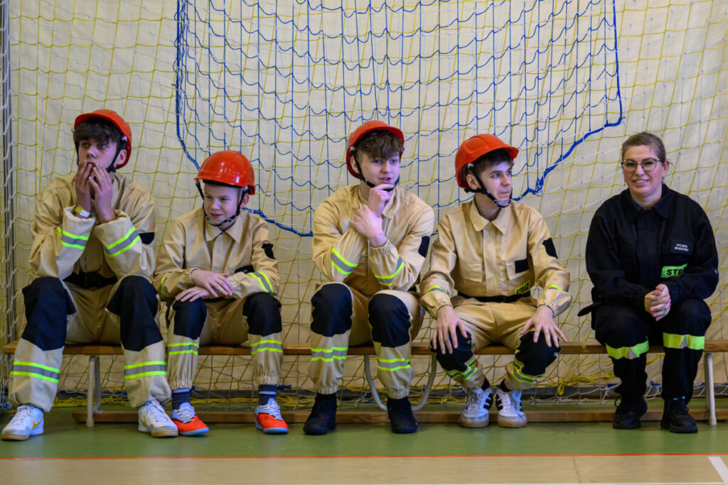 Pięć osób siedzi na ławce w stroju strażackim, cztery z czerwonymi kaskami. Tło stanowi siatka sportowa w żółto-niebieskich kolorach. Podłoga jest zielono-kremowa.
