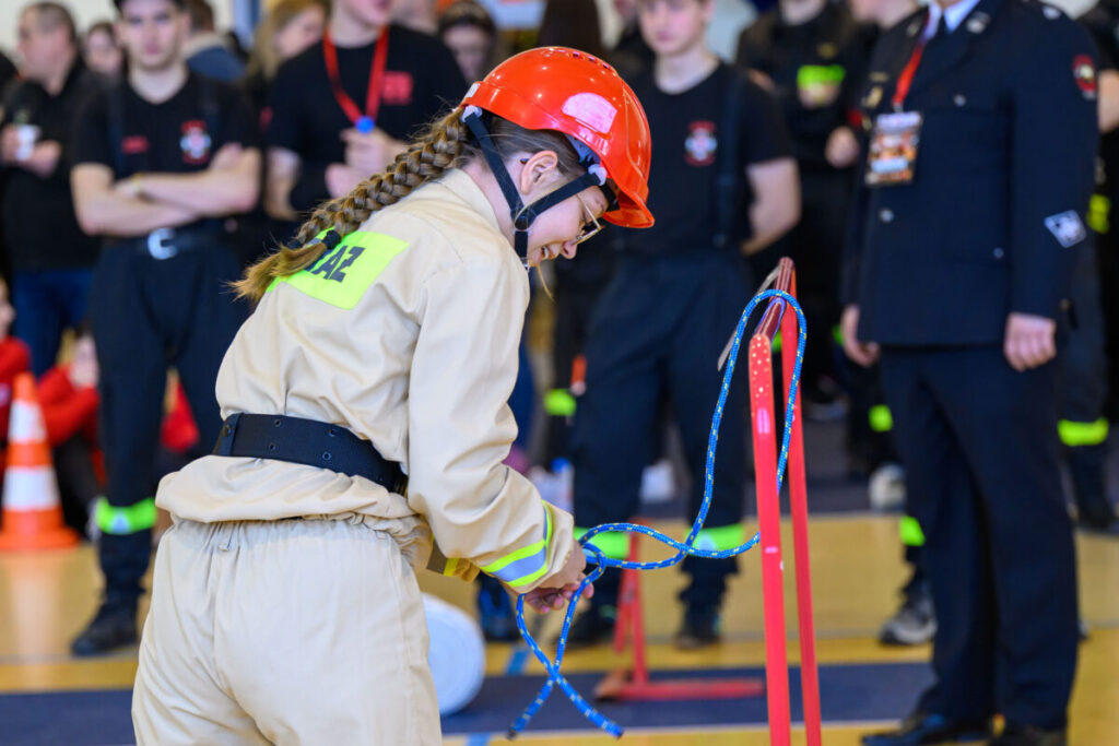 Młoda kobieta w stroju strażackim z czerwonym kaskiem wiąże linę. Tło niebieskawe, wokół stoją inne osoby w czerni. Na kurtce widoczny napis.