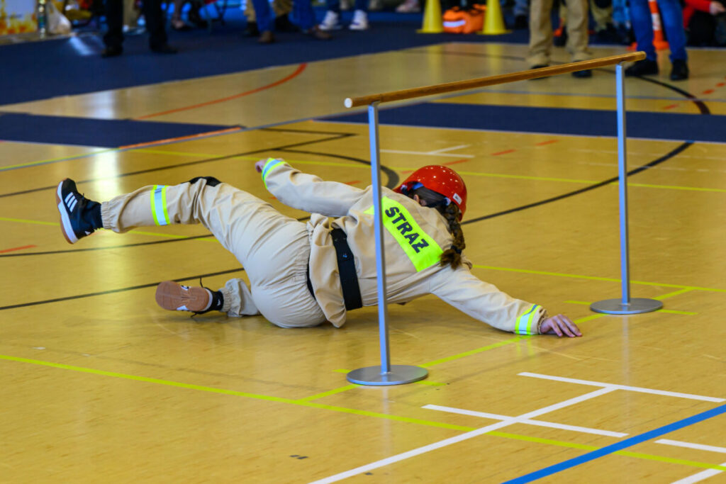 Osoba w stroju strażaka z hełmem czołga się po jasnym parkiecie, pod przeszkodą. Na plecach ma napis STRAŻ. Tło ukazuje ludzi w hali.