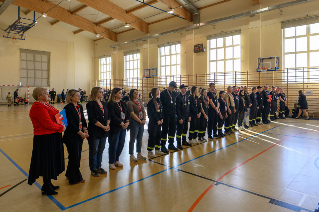 Grupa ludzi w uniformach stoi w szeregu w jasnej sali gimnastycznej. Drewniane belki na suficie. Tło stanowią okna i kosze do koszykówki.