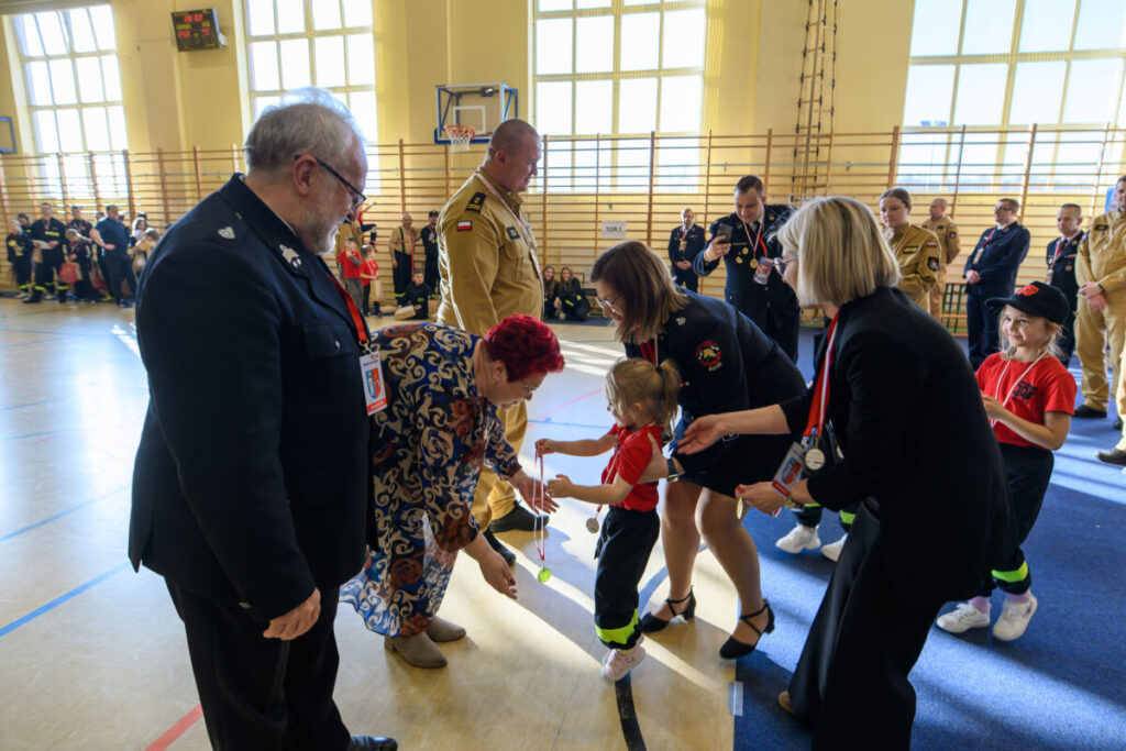 Grupa dorosłych i dzieci w sali gimnastycznej odbierających medale. Dziecko w czerwonej koszulce otrzymuje medal od kobiety. W tle zebrani ludzie i drabinki gimnastyczne.