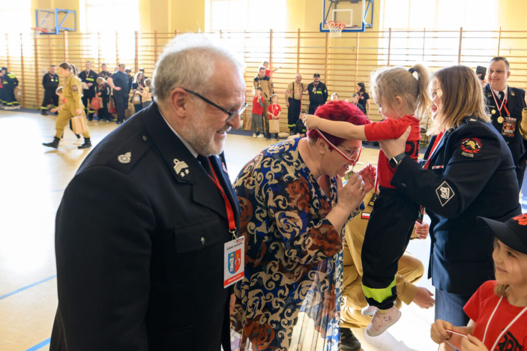 Na zdjęciu trwa wydarzenie w hali sportowej. W centrum są uśmiechnięci ludzie, dziecko w czerwonej koszulce. Tło jest jasne, widoczni strażacy na boku.
