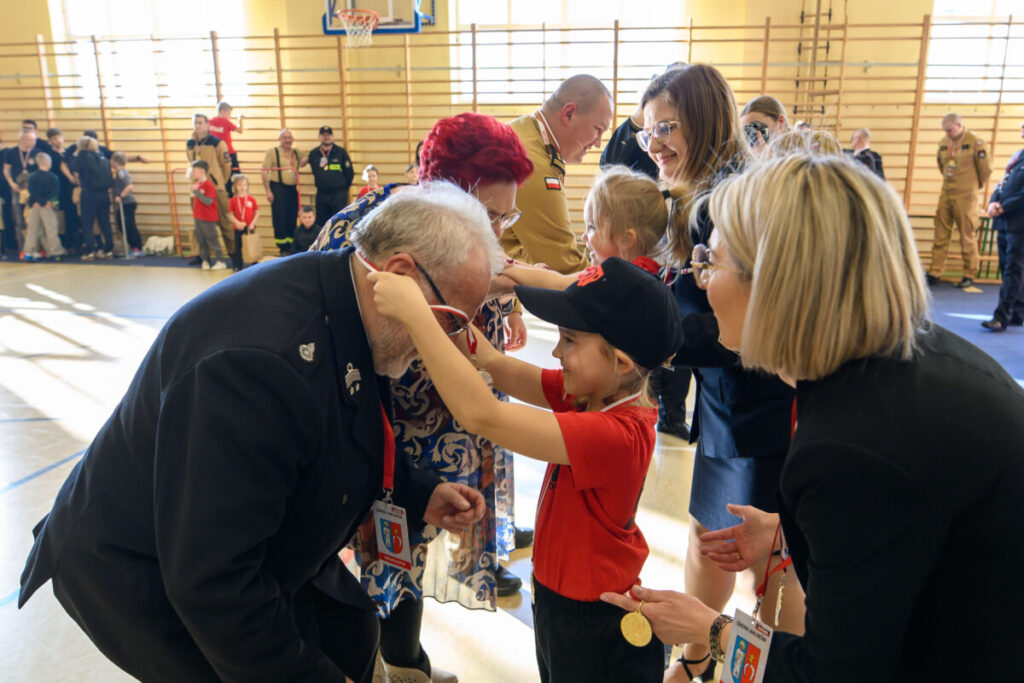 Dziecko w czerwieni zakłada medal starszemu mężczyźnie. Tło: sala gimnastyczna, jasne światło. Obecni: ludzie w różnych uniformach.