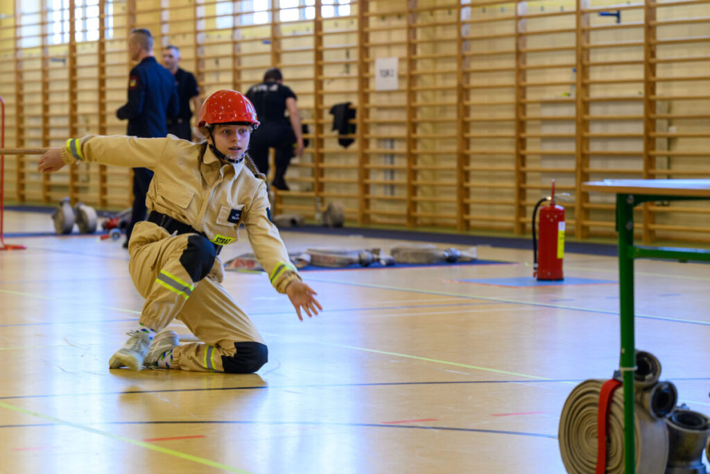 Osoba w kremowym stroju strażackim klęka na parkiecie sali gimnastycznej. Ma na sobie czerwony kask, w tle są ludzie i sprzęt. Podłoga oznaczona jest kolorowymi liniami.