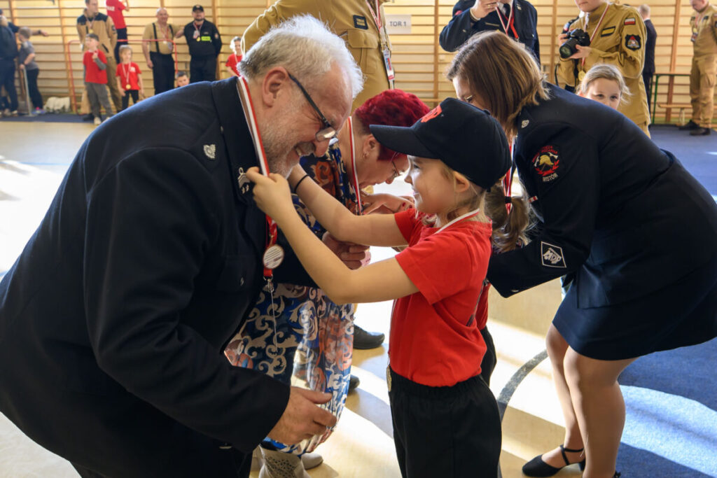 Na zdjęciu dziecko w czerwonym stroju zakłada medal starszemu mężczyźnie w ciemnym mundurze. W tle inni dorośli i dzieci na hali gimnastycznej. Wydarzenie jest uroczyste, dominują kolory czerwony i granatowy.