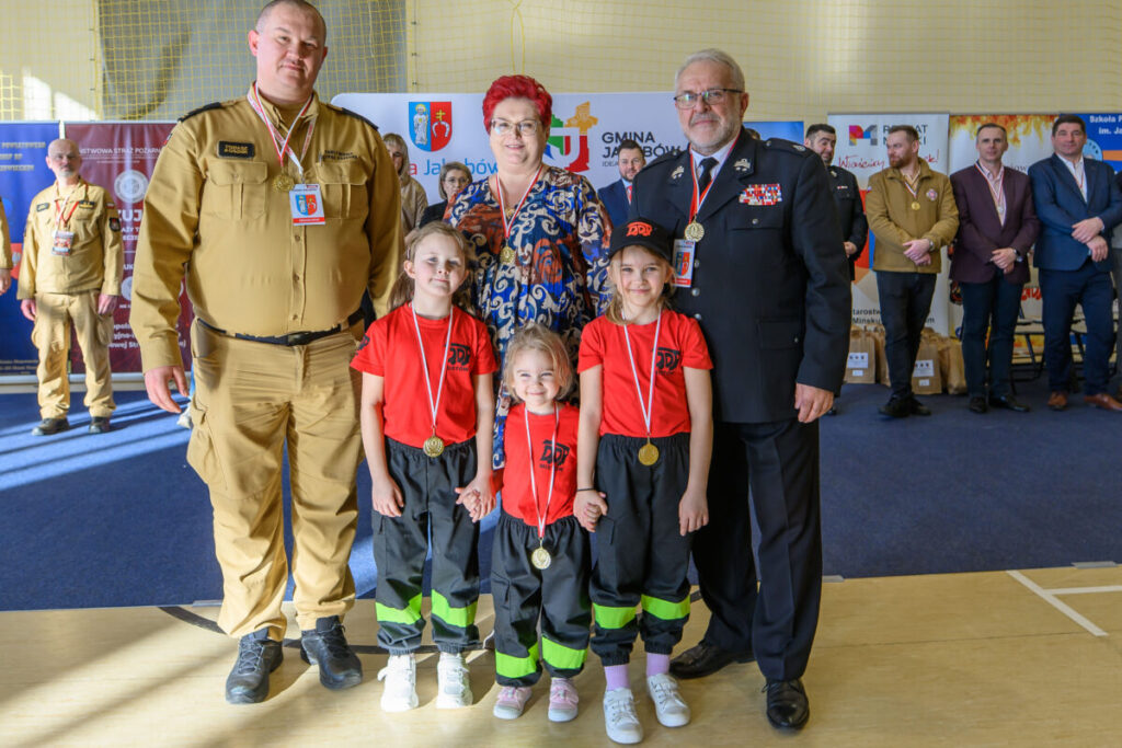 Grupa ludzi na zdjęciu to strażacy i dzieci w czerwono-czarnych strojach. W tle widać banery z kolorowymi logo. Scena oświetlona naturalnym światłem.