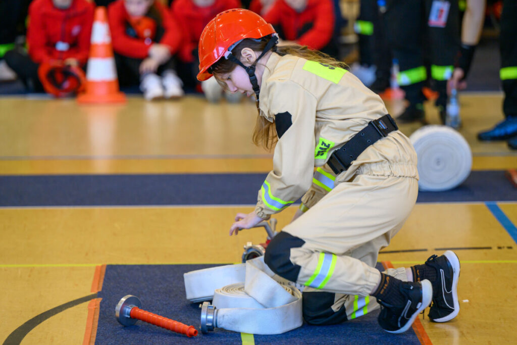 Dziewczynka w beżowym stroju strażackim i czerwonym kasku rozwija wąż na hali sportowej. W tle osoby w czerwonych ubraniach. Podłoga jest żółto-niebieska.