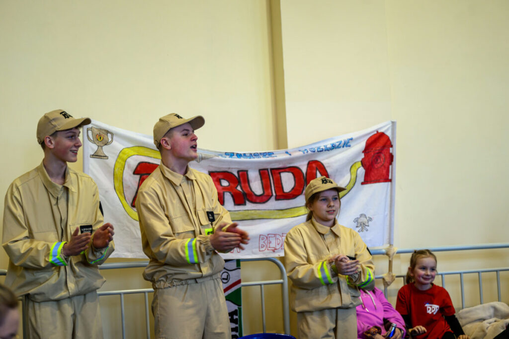 Trzy osoby w beżowych uniformach stoją przed kolorowym transparentem z napisem RUDA. W tle jasna ściana. Obok siedzi dwójka dzieci.