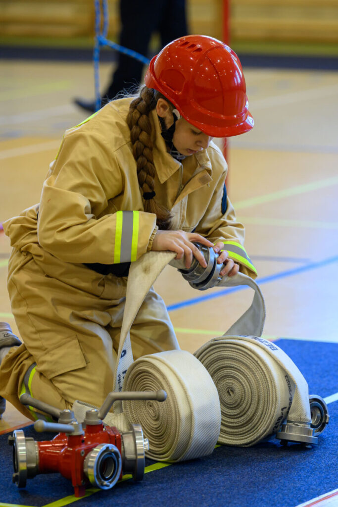 Osoba w stroju strażaka z czerwoną kaską układa wąż strażacki. Otoczenie to jasna sala z niebieskimi liniami na podłodze. Obok leży czerwona armatura wodna.