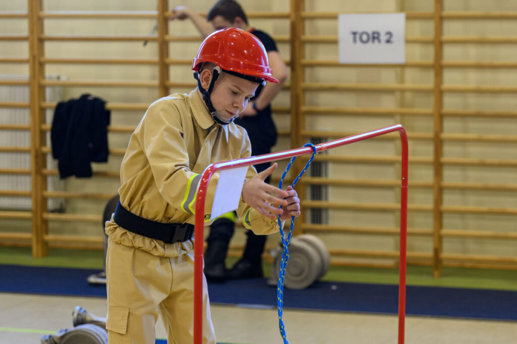 Chłopiec w czerwonym kasku i beżowym stroju strażackim wiąże węzeł na niebieskiej linie. W tle drewniana drabinka i znak TOR 2. Wnętrze sali gimnastycznej.