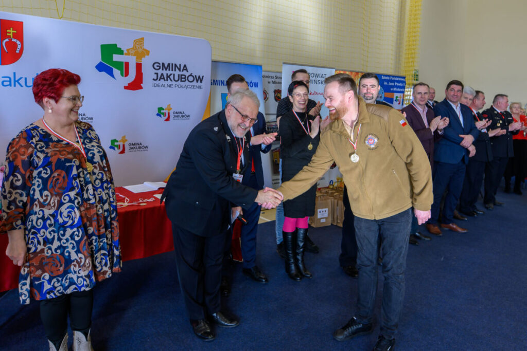 Mężczyzna w beżowej kurtce otrzymuje medal, ściskając dłoń starszego pana. Tło z logo gminy Jakubów. Grupa ludzi klaszcze.