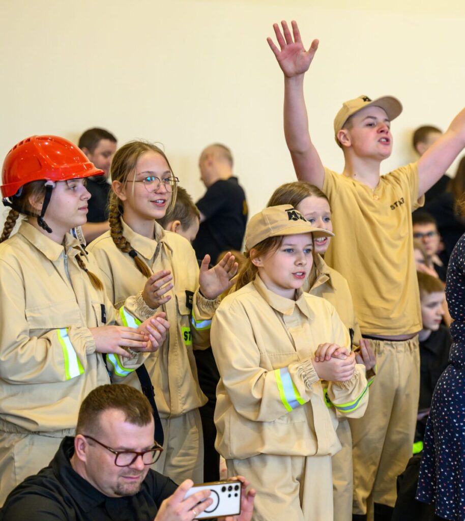Grupa dzieci w jasnych mundurach, jedna w czerwonym kasku, entuzjastycznie uczestniczy w wydarzeniu. Tło jest jasne, a dookoła widać innych uczestników. Na dole mężczyzna robi zdjęcie telefonem.