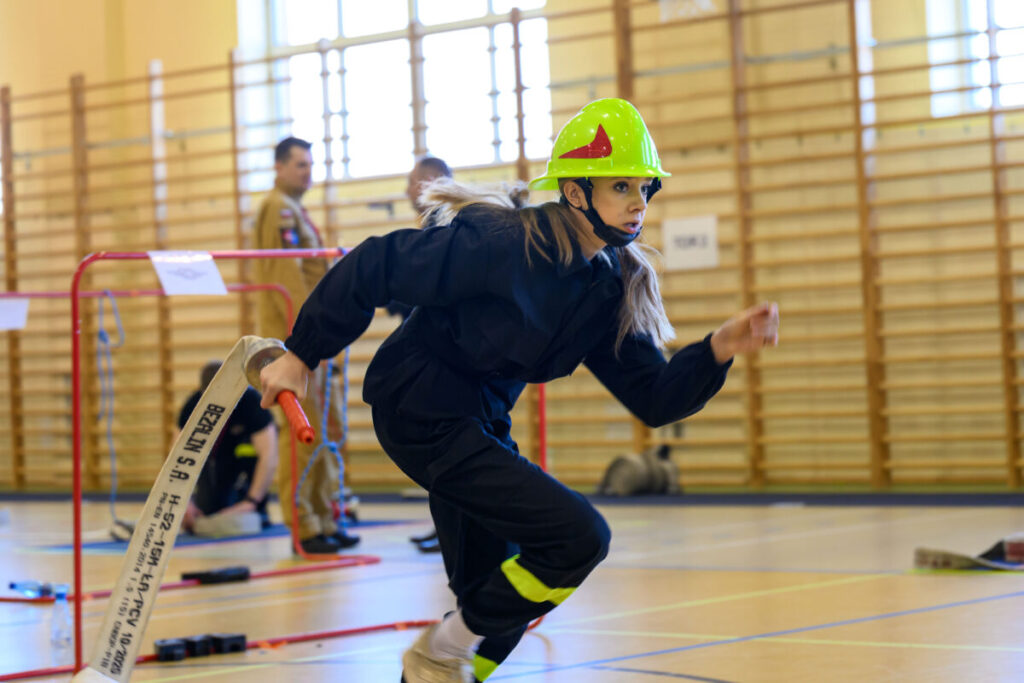 Młoda osoba w czarnym stroju strażackim i zielonym kasku biegnie z wężem strażackim na sali gimnastycznej. Tło to drewniane drabinki gimnastyczne i rozmyta postać. Pomieszczenie jest dobrze oświetlone przez