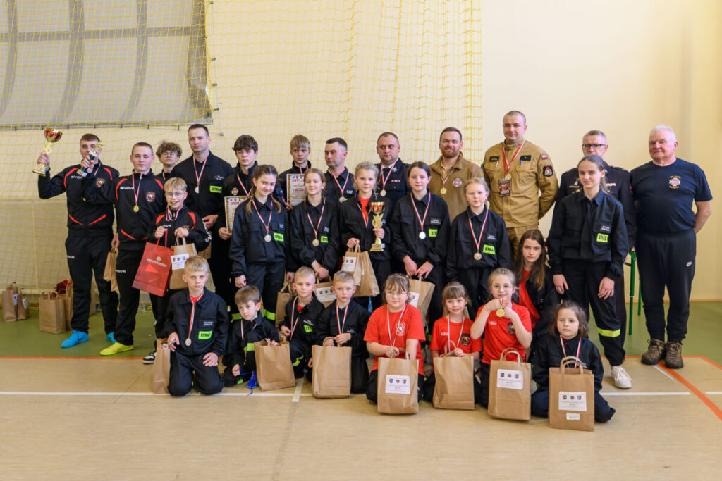 Grupa dzieci i dorosłych w czarnych oraz beżowych strojach strażackich pozuje z pucharami i medalami. Na podłodze leżą papierowe torby z upominkami. Tło stanowi jasna ściana i żółta siatka sportowa.