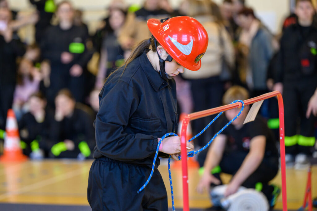 Młoda osoba w czerwonym hełmie wiąże węzeł na niebieskiej linie, ubrana na czarno. Tło rozmyte, widownia w ciemnych kolorach.