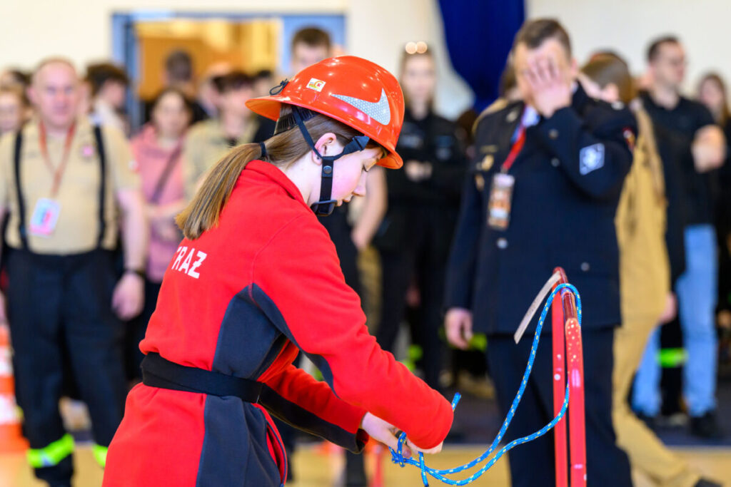 Osoba w czerwonym stroju strażackim wiąże niebieską linę. W tle ludzie w mundurach. Jasne pomieszczenie.