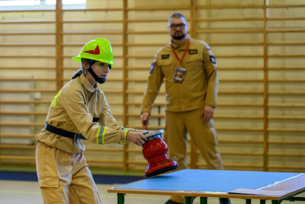 Młoda osoba w kasku manipuluje czerwoną maszyną na stole. Tło to sala gimnastyczna z drewnianymi poręczami. Osoba w mundurze obserwuje.