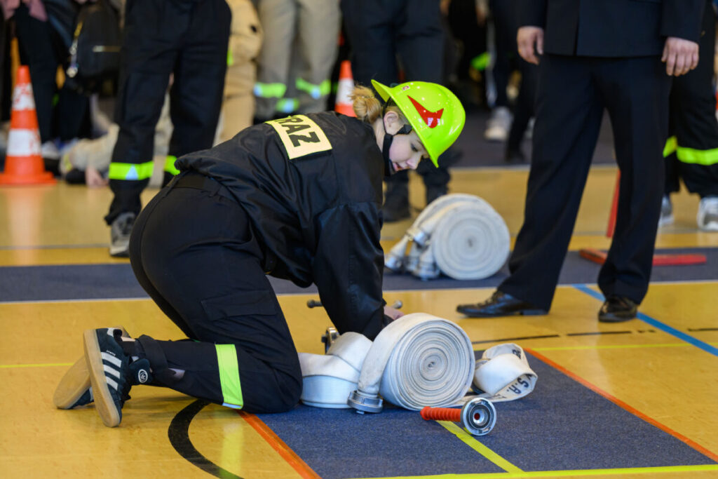 Młoda osoba w stroju strażackim na hali sportowej rozwija wąż. Ma na sobie kask w jaskrawym odcieniu. Inni strażacy widoczni w tle.