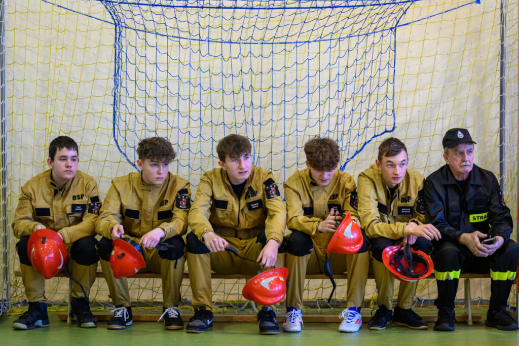 Sześciu młodych ludzi w żółtych mundurach z czerwonymi kaskami siedzi przed siatką w hali. Tło w barwach neutralnych. Obok nich siedzi starszy mężczyzna w czarnym uniformie.