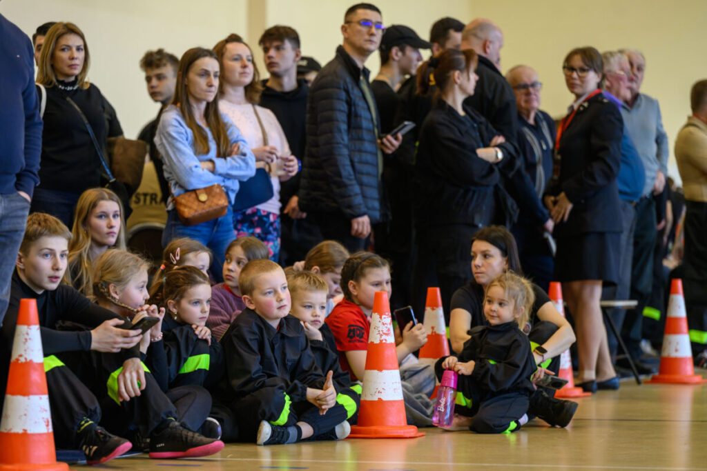 Grupa dzieci i dorosłych obserwuje wydarzenie w hali. Przeważają ciemne stroje, z widocznymi żółtymi elementami. Na podłodze stoją pomarańczowe pachołki.