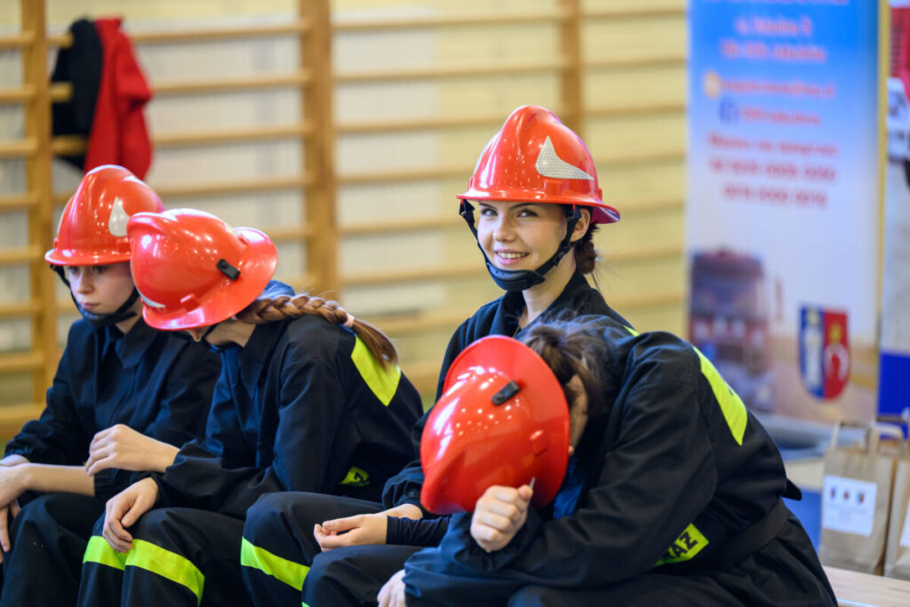 Cztery osoby w strażackich hełmach siedzą, jedna uśmiechnięta. Czerwone hełmy, czarne mundury. W tle drewniana drabinka i plakat.