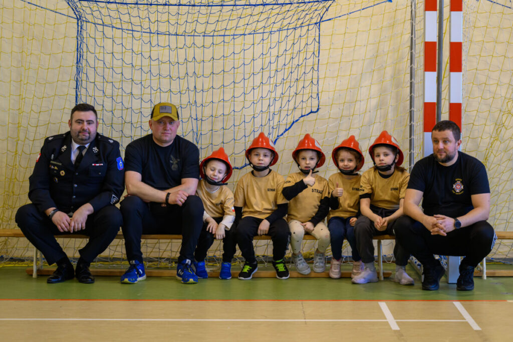 Grupa dzieci w czerwonych kaskach strażackich siedzi na ławce z trzema mężczyznami. Tło ma siatkę i żółtą ścianę. Dzieci mają beżowe koszulki z napisem STRAŻ.
