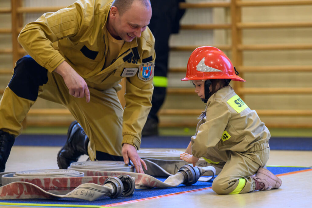 Strażak i dziecko w strojach strażackich układają wąż w sali gimnastycznej. Dziecko w czerwonym hełmie i beżowym stroju. W tle drabinki gimnastyczne.