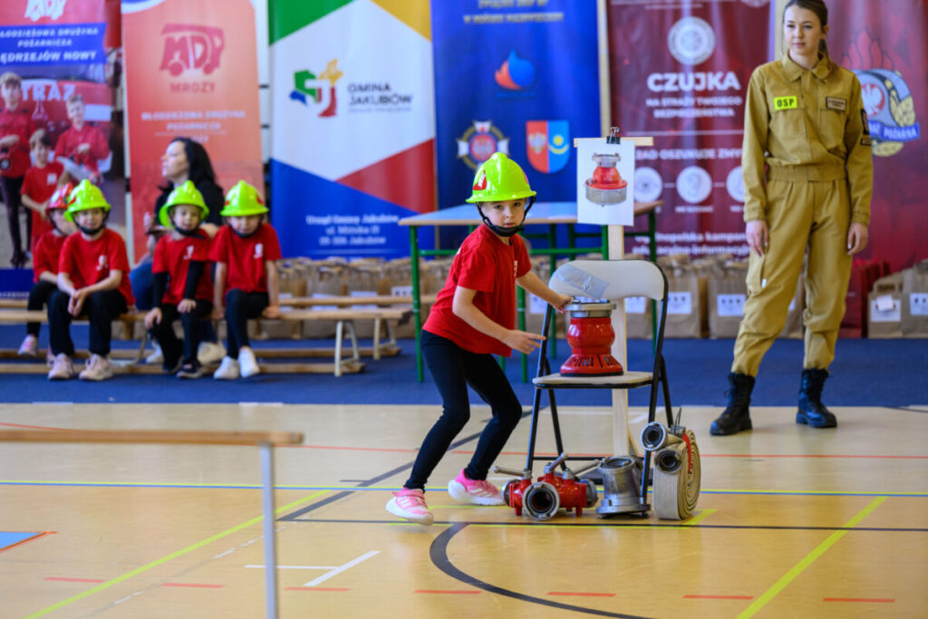 Dziecko w zielonym hełmie i czerwonej koszulce przy sprzęcie strażackim. W tle kolorowe bannery. Boisko sportowe.