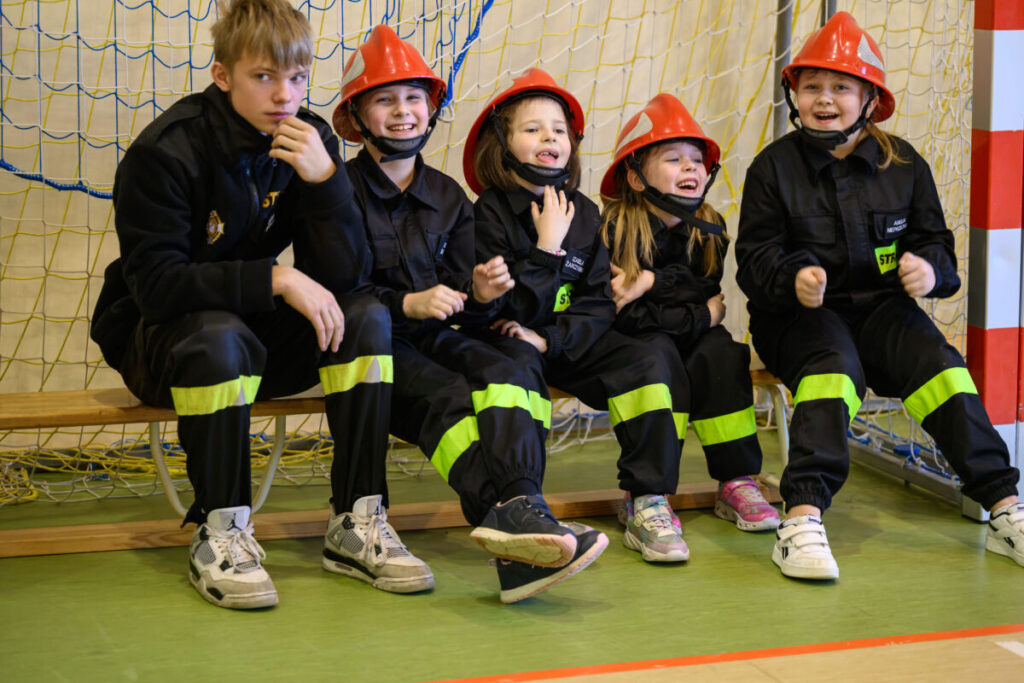 Czworo dzieci w czarnych strojach strażackich i czerwonych kaskach siedzi na ławce w sali gimnastycznej. Uśmiechają się, w tle niebieskie siatki. Podłoga jest zielona, obok stoi starszy chłopiec.