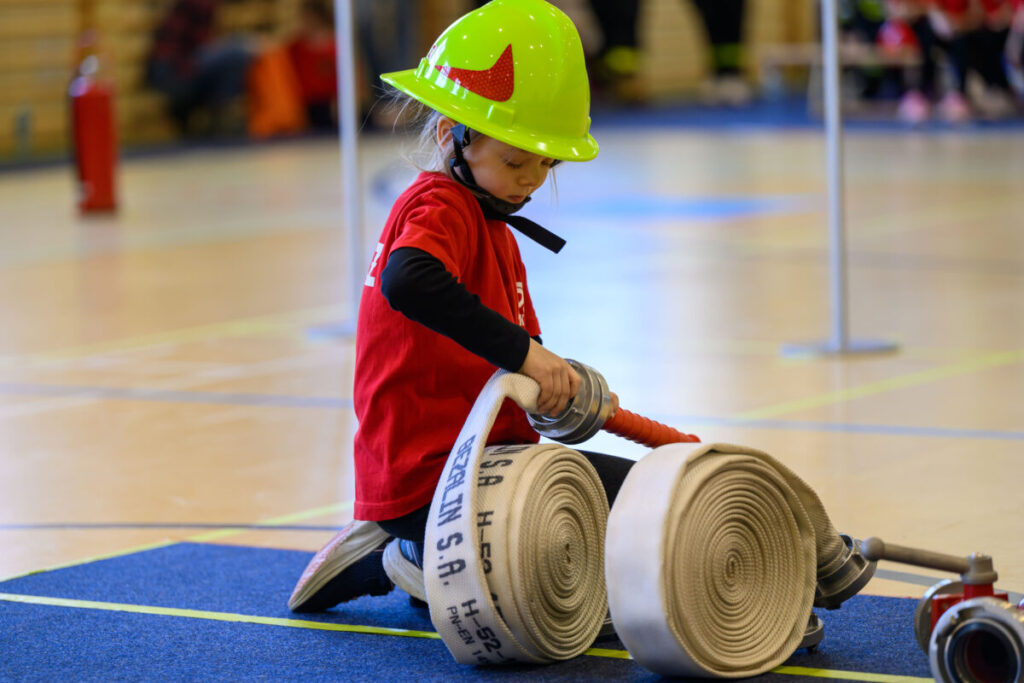 Na zdjęciu dziecko w kolorowym kasku, trzymające zwinięty biały wąż strażacki. Ma na sobie czerwoną koszulkę, stoi na niebieskiej macie. W tle sala gimnastyczna.