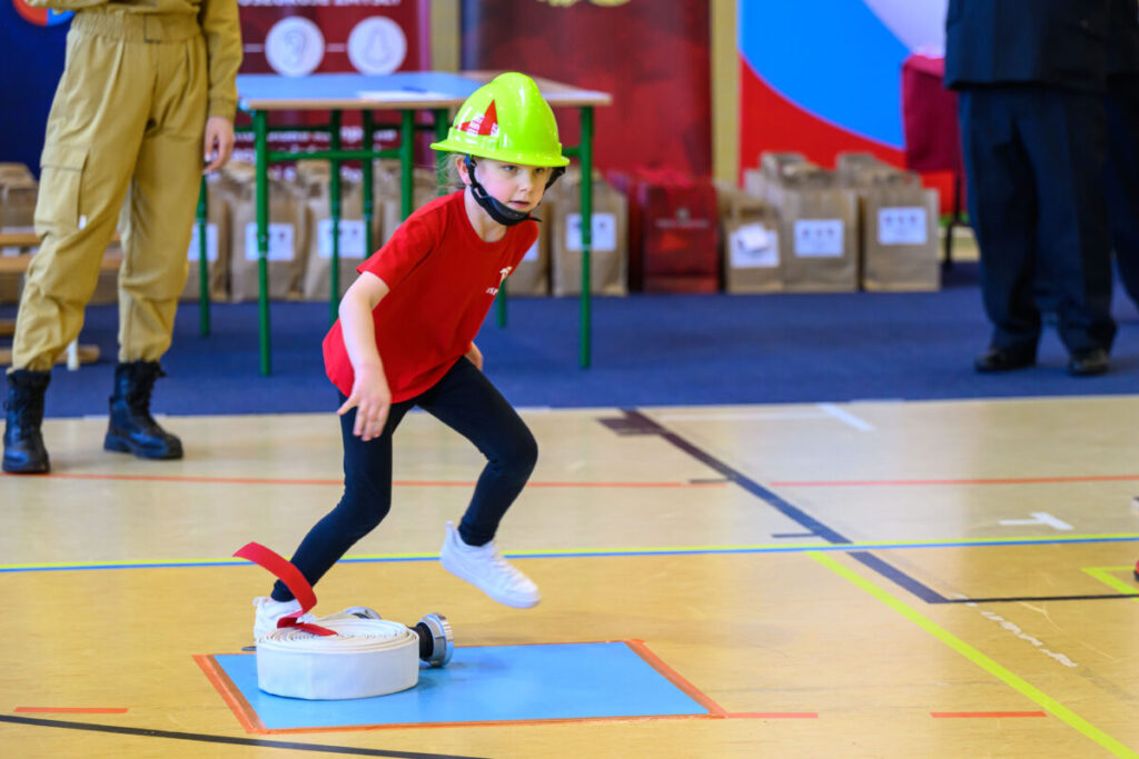 Dziecko w czerwonej koszulce i zielonym kasku bierze udział w zawodach strażackich. Na podłodze leży biały wąż. W tle stoją osoby i torby na niebiesko-czerwonym tle.