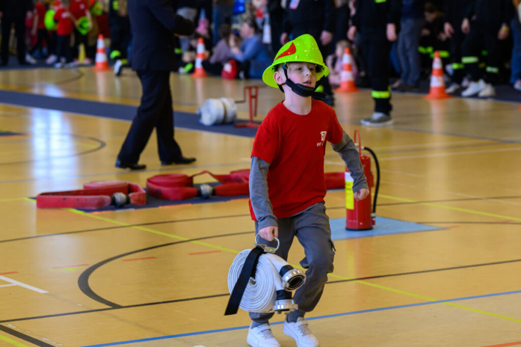 Młody chłopiec w zielonym hełmie i czerwonej koszulce niesie wąż strażacki. Tło to sala gimnastyczna z kolorowymi przeszkodami. W tle ludzie i pachołki.