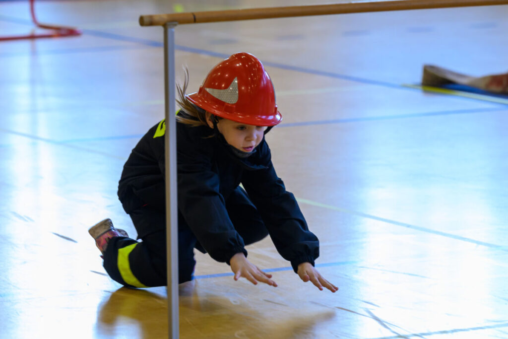 Dziecko w czerwonym kasku strażackim kuca na jasnej podłodze sali gimnastycznej. Tło jest niebieskawe. Obok widoczny jest tyczek.