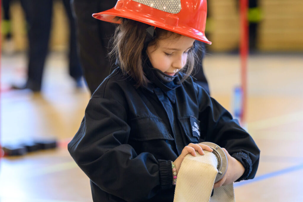 Dziecko w czerwonym hełmie strażackim i czarnym stroju trzyma wąż strażacki. Tło jest rozmyte w jasnych kolorach.