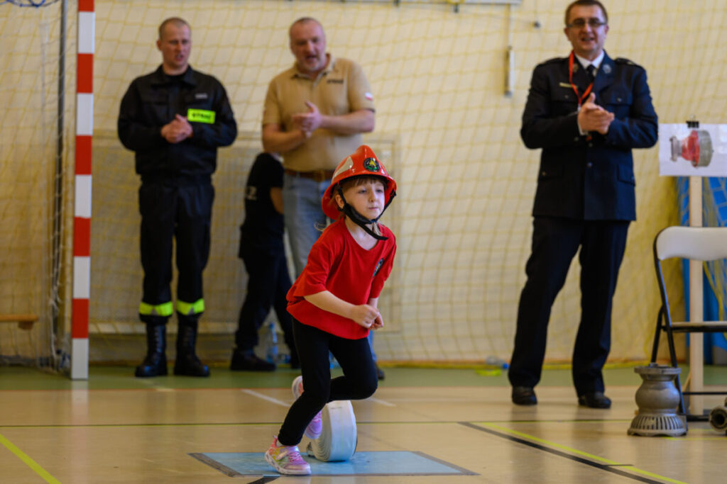 Na zdjęciu dziecko w czerwonym kasku i koszulce biegnie po hali sportowej. W tle siatka i kilku dorosłych, w tym strażak. Obok dziecka leży zwinięty wąż strażacki.