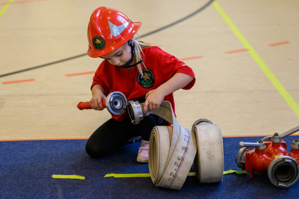 Dziecko w czerwonym hełmie i koszulce siedzi na kolanach, trzymając wąż strażacki na niebieskim dywanie. Tło jasne, beżowe. W pobliżu leży rozwinięty wąż.