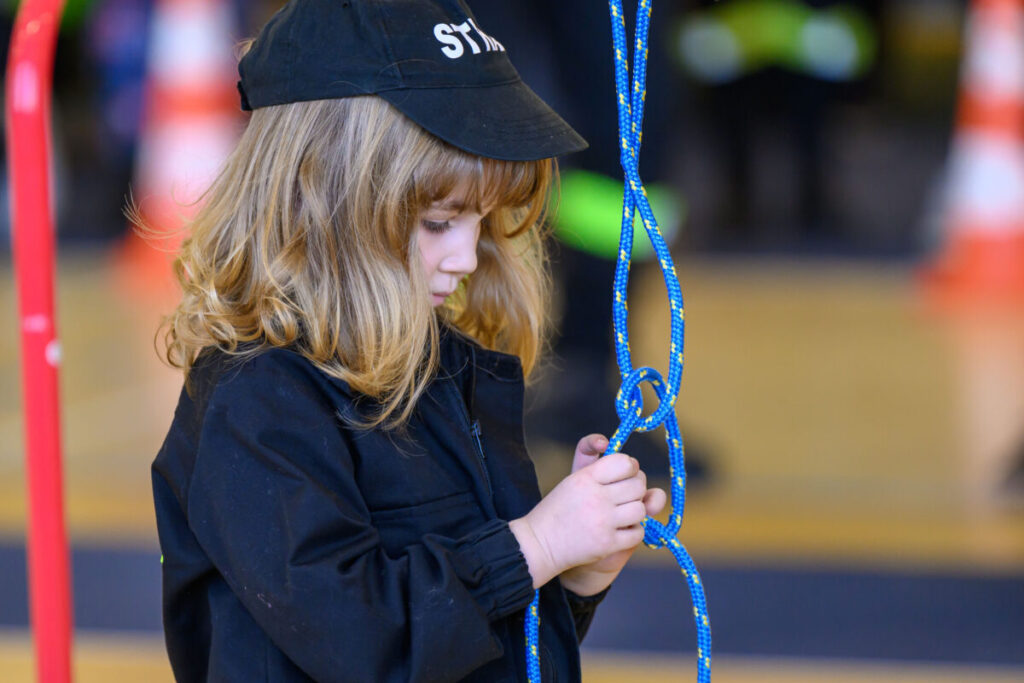 Małe dziecko w czarnej czapce i bluzie wiąże niebieską linę. Tło jest rozmyte z pomarańczowymi elementami. Skupiona mina dziecka.