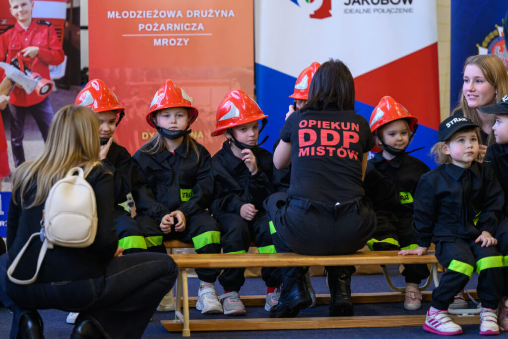 Grupa dzieci w czerwonych kaskach strażackich siedzi na ławce. Noszą czarne uniformy z zielonymi paskami. W tle są kolorowe banery na ścianie.
