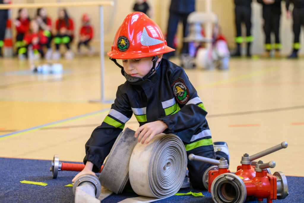 Na zdjęciu jest dziecko w stroju strażackim z czerwonym hełmem. Siedzi na podłodze, zwija wąż strażacki. W tle widać rozmazane sylwetki ludzi w czerwonych uniformach.