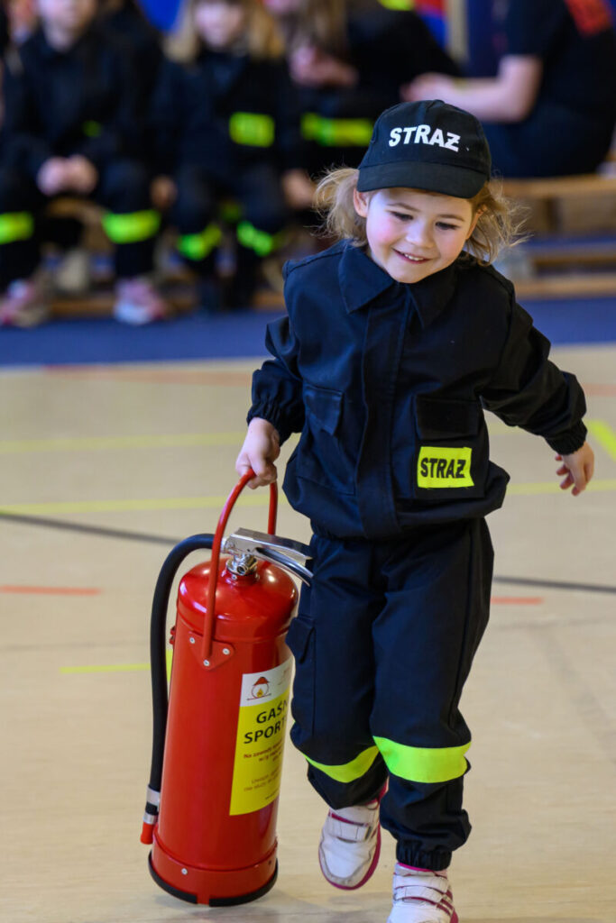 Małe dziecko w czarnym mundurze z napisem STRAŻ niesie czerwony gaśnicę. Tło to sala wewnętrzna z siedzącymi dziećmi. Dominują ciemne kolory i żółte akcenty.