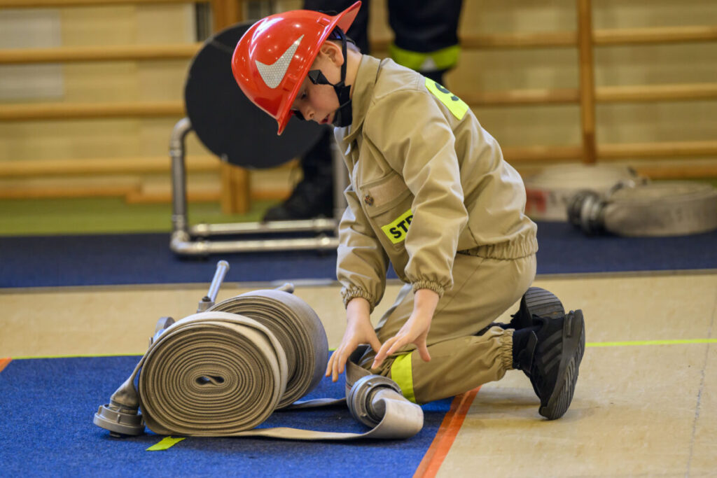 Młody strażak w czerwonym hełmie zwija wąż pożarniczy na niebieskiej macie. W tle drewniane drabinki gimnastyczne. Dominują beże i czerwienie.