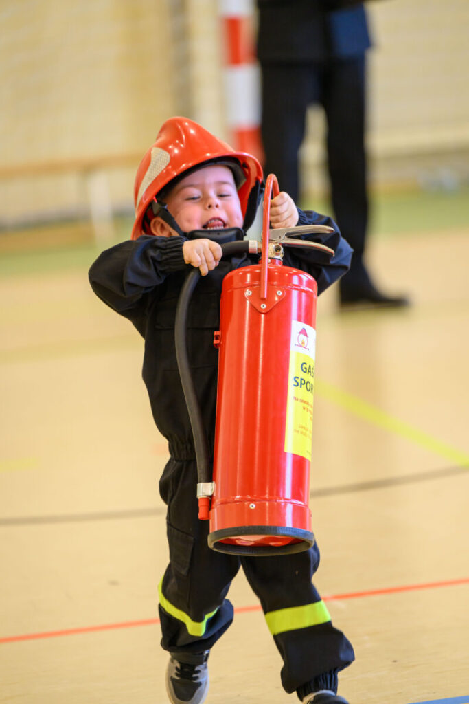 Na zdjęciu dziecko z hełmem strażackim trzyma gaśnicę. Ma na sobie czarny strój z odblaskowymi elementami. W tle widoczna jest sala.