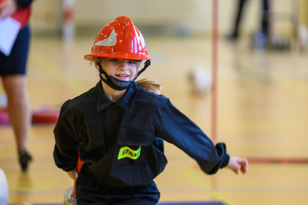 Dziewczynka w czerwonym hełmie strażackim i czarnym stroju biegnie po hali. Jasne tło, niewyraźne postacie w tle. Uśmiechnięta, z napisem STRAŻ.