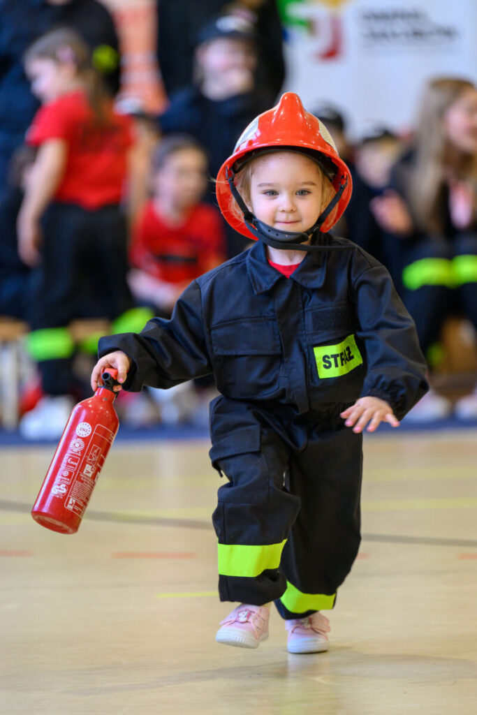 Dziewczynka w stroju strażaka z czerwonym kaskiem i gaśnicą. Czarny strój z zielonym elementem. Tło z rozmytymi postaciami.