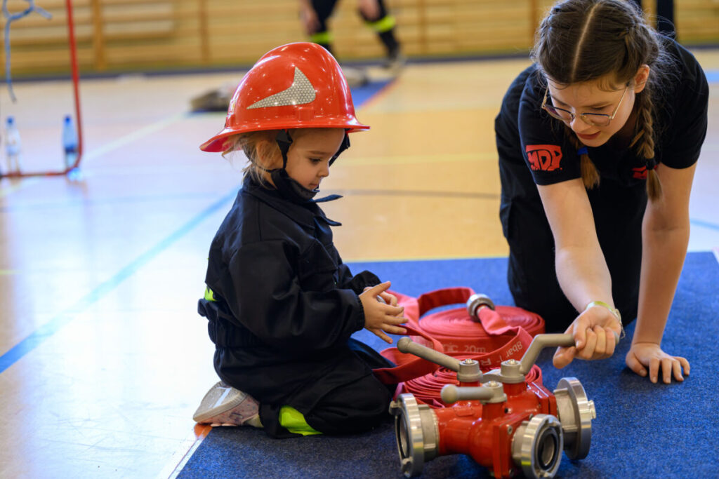 Małe dziecko w czerwonym hełmie strażackim i czarnym kombinezonie. Obok niego starsze dziecko wskazuje coś na wężu strażackim. Podłoga jest jasna z niebieskim dywanikiem.