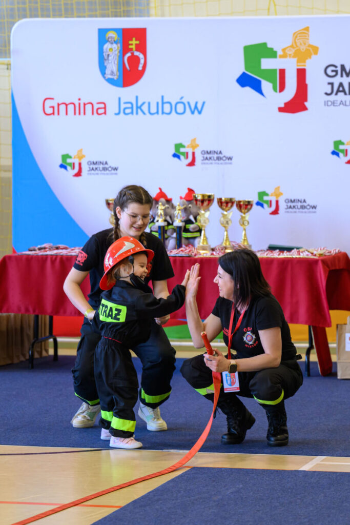 Na zdjęciu jest dziecko w stroju strażaka, przybijające piątkę z instruktorką. W tle są puchary na czerwonym stole i logo gminy Jakubów na banerze. Kolory dominujące to czerwony, czarny i niebieski.