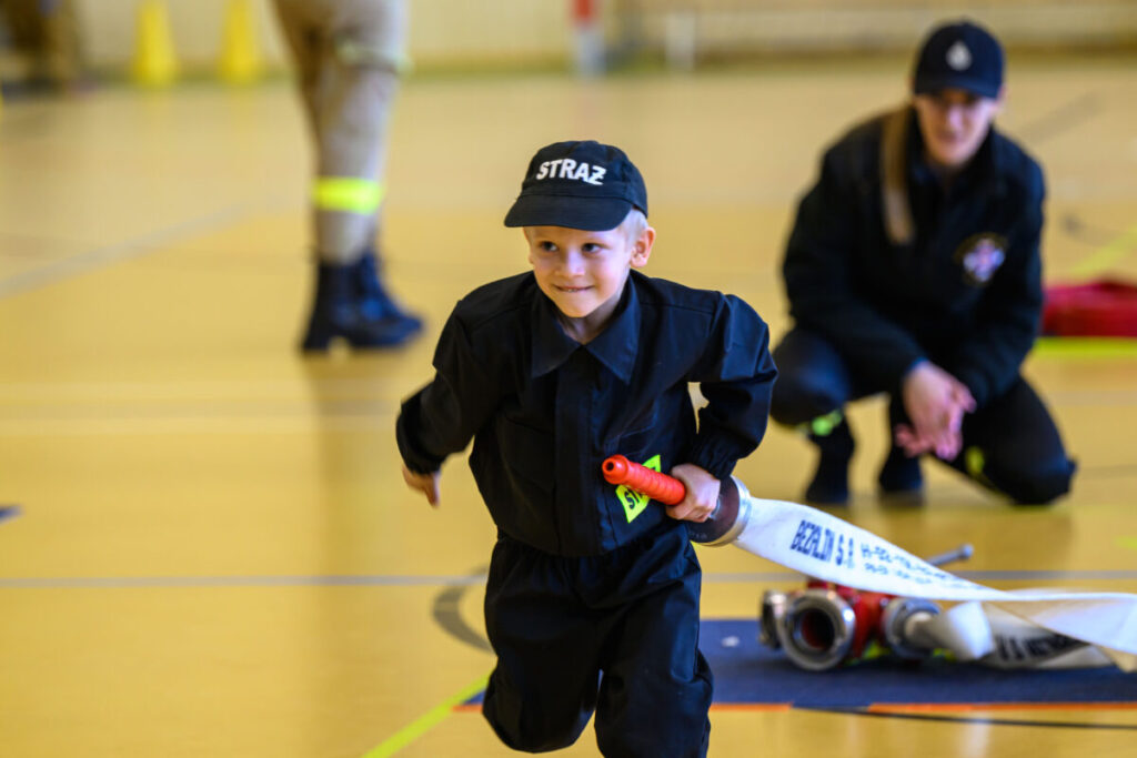 Młody chłopiec w stroju strażaka biegnie z wężem na jasnym tle. Za nim klęczy dorosła postać. Dynamiczna scena w hali sportowej.