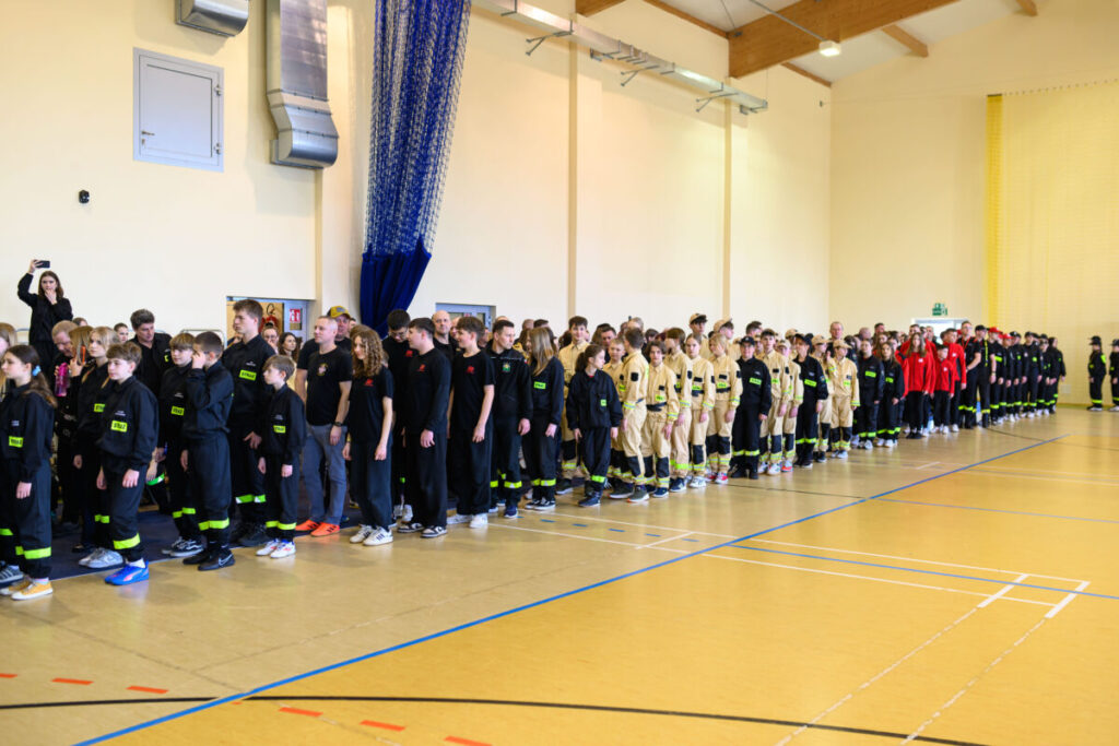 Grupa młodych ludzi w mundurach strażackich stoi w rzędach na sali gimnastycznej. Mundury są czarne, czerwone i beżowe z żółtymi akcentami. W tle jasne ściany i niebieska zasłona.