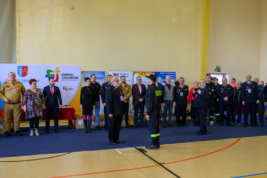 Grupa ludzi w mundurach i garniturach stoi w dużej sali. Tło z logo i banerami w jasnych kolorach. Mężczyzna w mundurze strażackim stoi na środku.