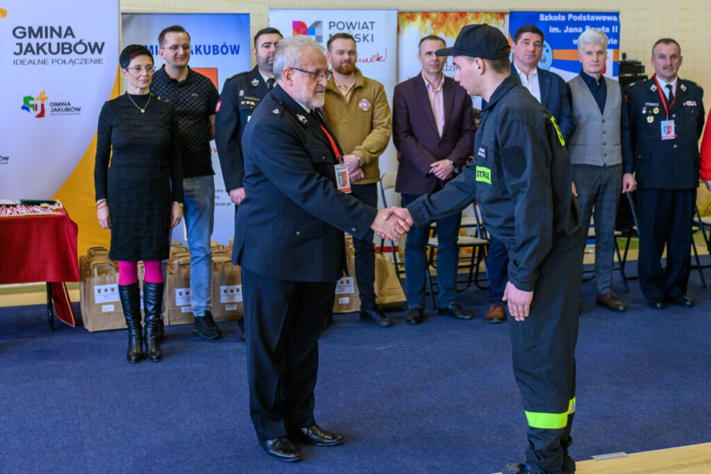 Na zdjęciu strażacy w ciemnych mundurach w uścisku dłoni. Tło to kolorowe bannery i osoby w eleganckich strojach. Dominują odcienie niebieskiego i żółtego.